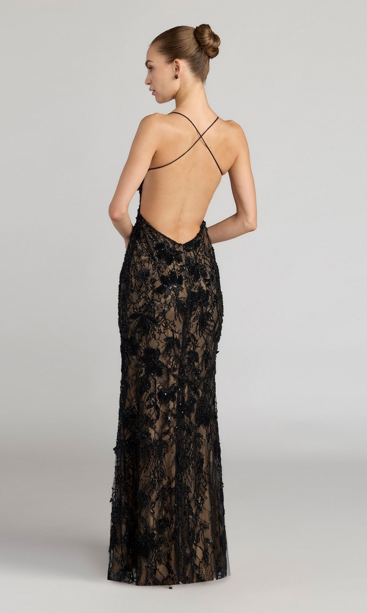 A woman models the Mac Duggal 68892 long formal dress—a black, floor-length column gown with beaded lace embroidery, low-cut sleeveless open back, and crisscross straps—standing turned away from the camera.